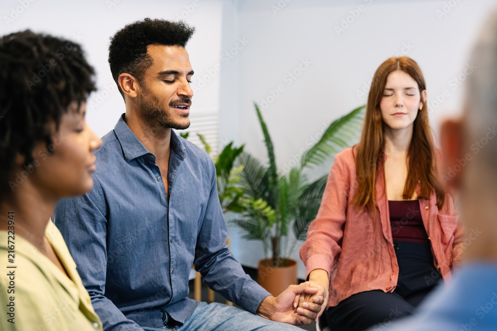 Obraz premium Group of people sitting in a circle, holding hands, and meditating with closed eyes, finding emotional support and community in a therapy session for improved mental health and wellness