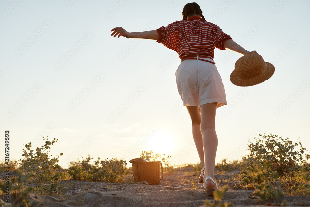 Fototapeta premium Young woman in a striped top and white shorts walks away at sunset, holding a wide hat, strolling along a dusty path as warm light glows on her silhouette.