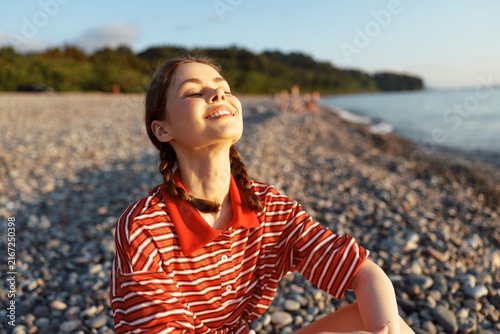 Bild auf Leinwand Young woman on a pebbled beach basks in warm sunlight, smiling with eyes closed, enjoying a casual summer moment