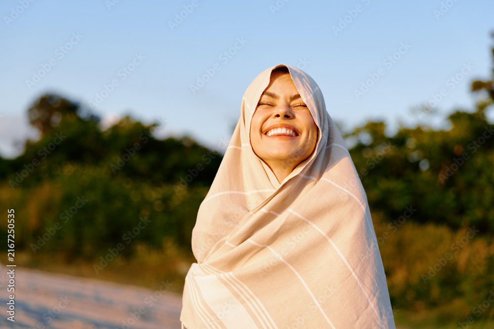 Fototapeta premium Smiling woman wrapped in a light shawl outdoors on a sunny day, enjoying a gentle breeze. Lifestyle portrait for wellness, travel, or nature themes.