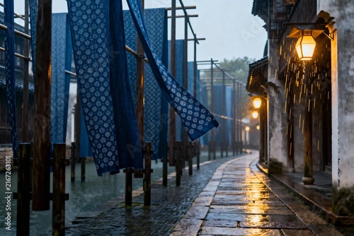 Wet stone pathway lined with blue indigo-dyed fabrics hanging on wooden poles beside traditional buildings with glowing lanterns at dusk