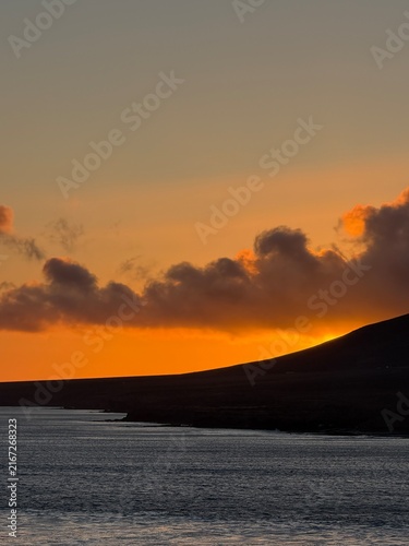 Canary Islands. Sunset view over the ocean with bright orange and yellow colors reflecting on the water, mountains and clouds framing the serene coastal landscape