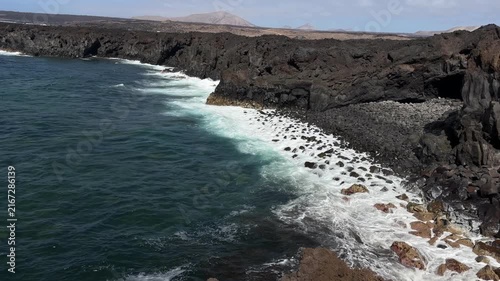 Canary Islands. Rocky coastline features dark volcanic cliffs with waves crashing against them, set against a clear blue sky and distant landforms in the background