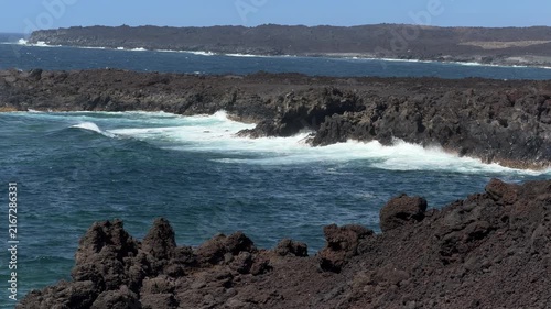 Canary Islands. Rocky coastline features dark volcanic cliffs with waves crashing against them, set against a clear blue sky and distant landforms in the background