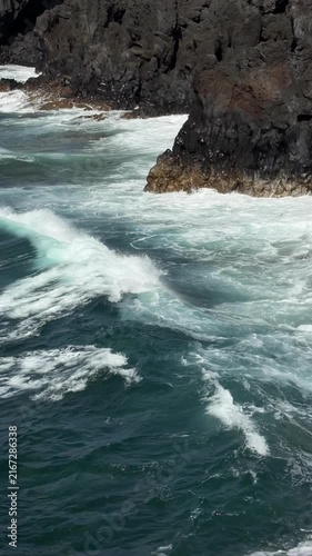 Canary Islands. Rocky coastline features dark volcanic cliffs with waves crashing against them, set against a clear blue sky and distant landforms in the background