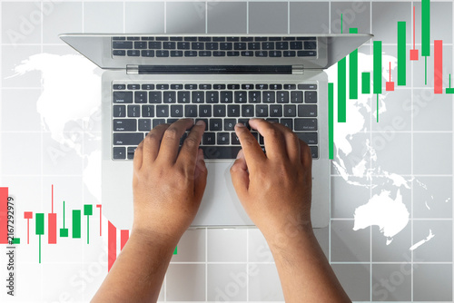 Top view of hands typing on a laptop with a digital overlay of red and green candlestick charts and a world map. Concept of global stock market trading, cryptocurrency investment, and finance.