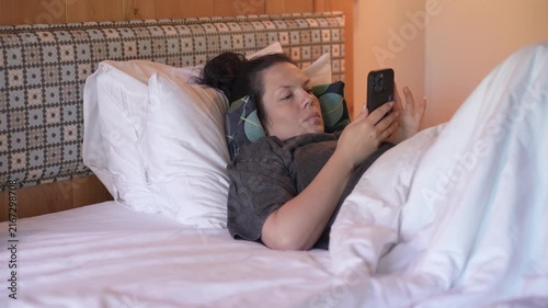 Woman is lying in bed covered with a blanket, texting on the phone. Girl under the blanket sends a message with his smart phone and smiles because he received an amusing message or love, and responds 