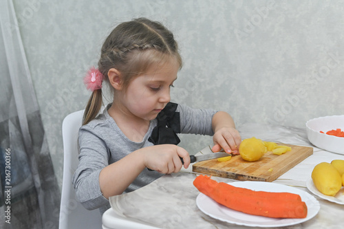 little girl at kitchen table demonstrates cooking education by cutting vegetables. Small kitchen helper cuts boiled potatoes, developing motor skills while participating in family cooking activities.
