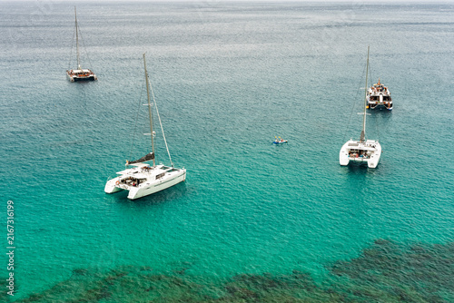 Aerial view of catamarans and sailboats anchored in turquoise water with kayak nearby.