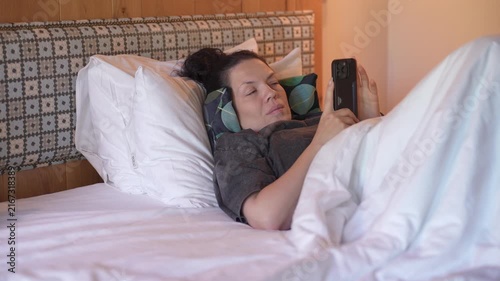 Woman is lying in bed covered with a blanket, texting on the phone. Girl under the blanket sends a message with his smart phone and smiles because he received an amusing message or love, and responds 