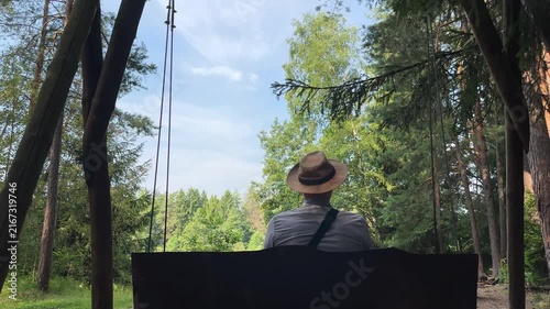 Man in straw hat swinging on wooden bench in summer forest view from behind. Male tourist relaxing outdoors and looking at green tree canopy in countryside park. Leisurely outdoor activity