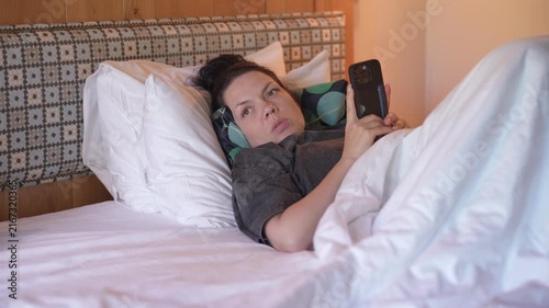 Woman is lying in bed covered with a blanket, texting on the phone. Girl under the blanket sends a message with his smart phone and smiles because he received an amusing message or love, and responds 