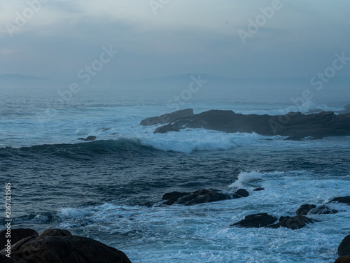 Choppy ocean waves with white foam crashing over dark rocks along a foggy, overcast coastline under a soft blue hazy sky.
