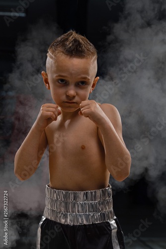 Close up shot of a determined young boy in boxing trunks holding up fists in defensive stance surrounded by dramatic studio smoke in an indoor fitness club.
