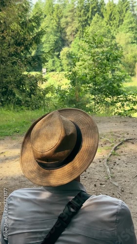 Man in hat sitting outdoors and admiring summer forest scenery view from back. Traveler enjoying tranquil day in green woodland park surrounded by tall pine trees. Peaceful outdoor leisure