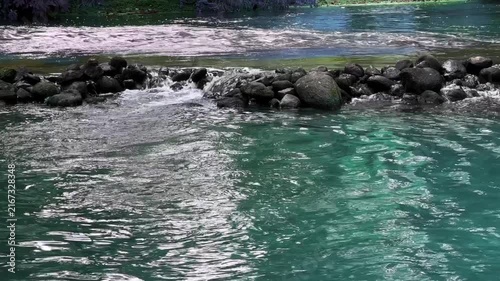 Clear turquoise blue and green water mixing at rocky river threshold. Close view of turbulent bubbling water stream flowing over river stones. Vibrant natural water texture and wave movement