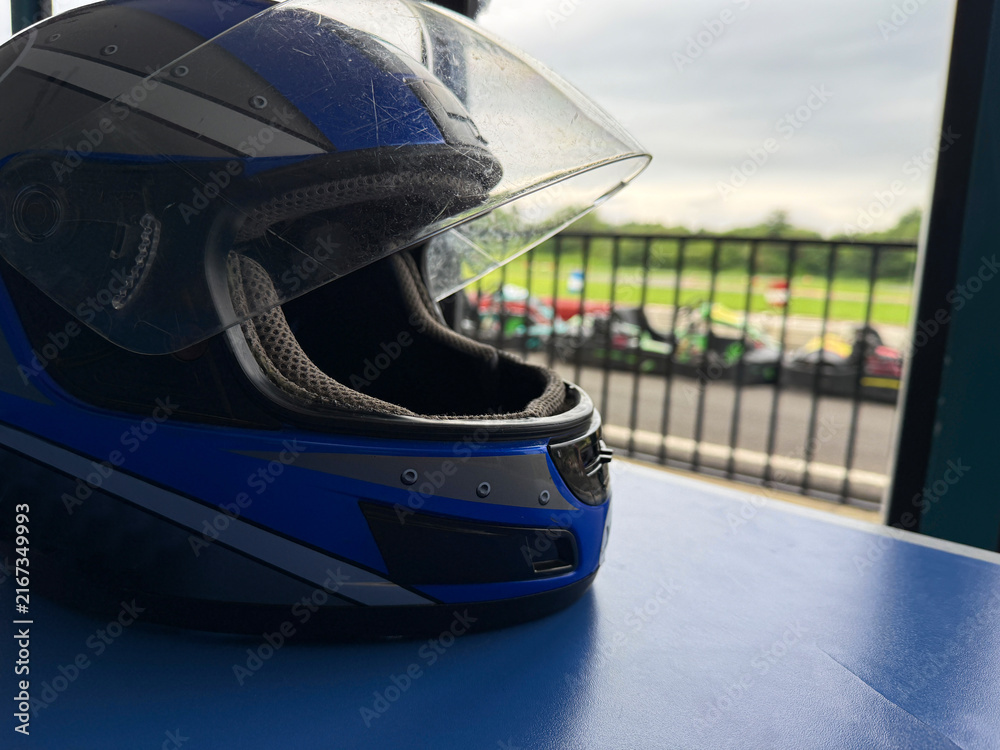 Fototapeta premium A blue racing helmet is placed on a table while go-karts are visible in the background.