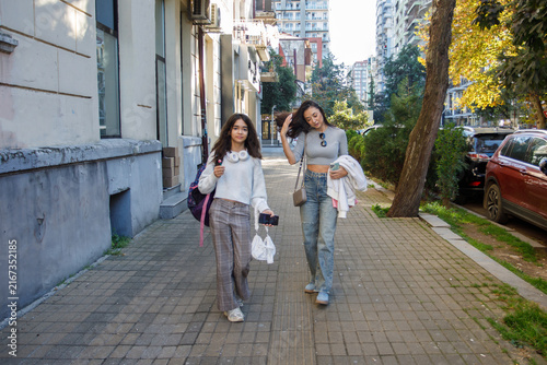 Two people walk on a city sidewalk in the afternoon with buildings and trees in the background