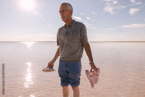Senior man in shallow pink salt lake water, holding children's pink clogs in both hands. Bright sunlight, calm water surface, blue sky with light clouds, copy space.