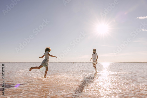 child running through shallow pink salt lake water toward mother under bright summer sun, family vacation, joyful outdoor travel scene with water splashes, tourism, travel agency, parenting