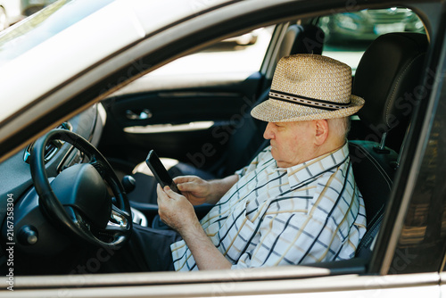 Older male driver checking his mobile phone inside the vehicle cabin outdoors.