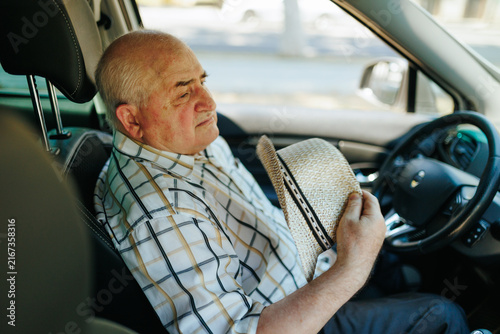 Older male driver takes off his hat while resting inside the car cabin outdoors.