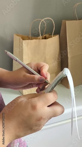 Woman writing a grocery shopping list in a notebook with paper bags of groceries in the background, shopping trip, food expenses and household planning concept.