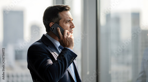 Professional man engaged in important phone call standing by large window overlooking cityscape in modern office environment
