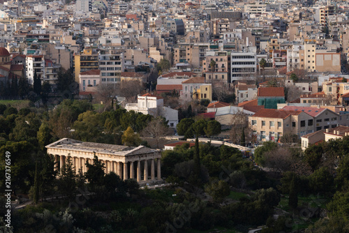 Aerial view of the well-preserved ancient Temple of Hephaestus nestled in the lush Ancient Agora park, contrasting with the dense, modern residential rooftops of Athens, Greece.