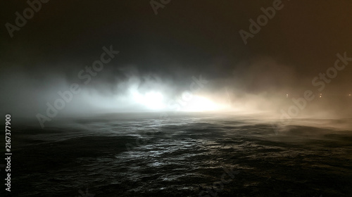 Konstfotografi Mysterious bright lights shine through thick fog over dark turbulent water at night