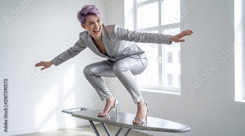 Cheerful woman in elegant silver suit surfing on ironing board in bright studio