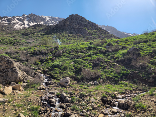 Quadro su tela Sun-drenched green hillside rises toward snow-capped peaks, thin smoke drifting skyward beside a flowing stream under a brilliant blue spring sky