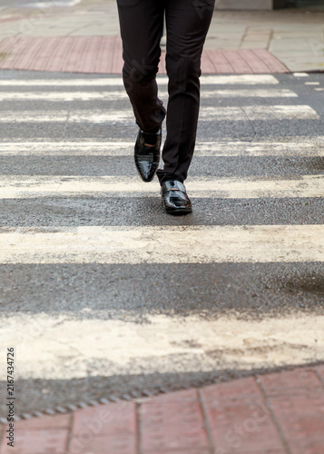 A person in formal black trousers and polished dress shoes walks across a pedestrian crosswalk in the city, suggesting a professional commute or business-related activity.