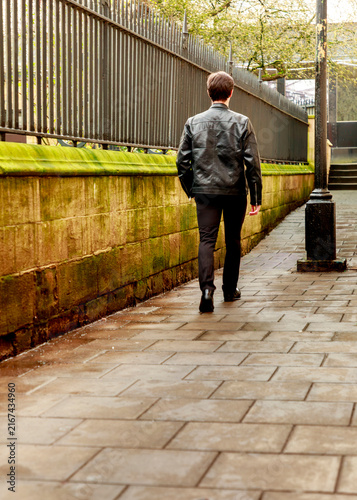 A man dressed in a black leather jacket walks away from the camera along a historic stone path beside a tall metal fence on a quiet day.