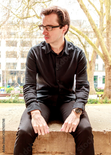 A young man wearing a black shirt and glasses poses thoughtfully while sitting on a stone ledge in a park, capturing a contemplative moment during an outdoor portrait session.