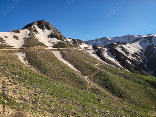 Canvas Print Snow-dusted alpine peak towers over vibrant emerald hillsides, where a winding trail weaves through spring meadows beneath a flawless azure sky