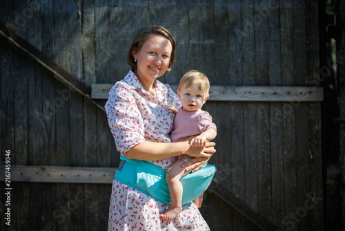 A woman holding a baby in a hip seat against a dark wooden wall