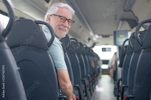 Senior adult man with grey hair and beard smiling while looking back from his seat on a public transport bus, enjoying independent travel and comfortable commuting