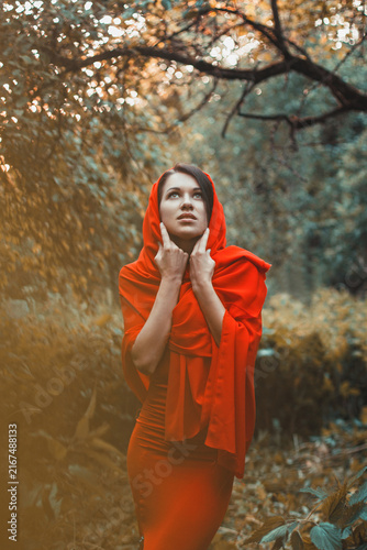 Konstfotografi woman in a vibrant red dress and hooded wrap stands in a sunlit forest, gazing upward with a thoughtful expression