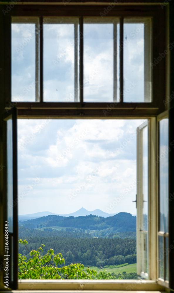 Fototapeta premium Scenic landscape view of Bezdez castle twin peaks framed by an open old wooden window looking out over green forests and hills under a cloudy summer sky.