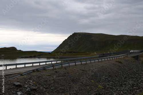 Photography A winding coastal road curves along green hills and a calm fjord under an overcast sky in Norway