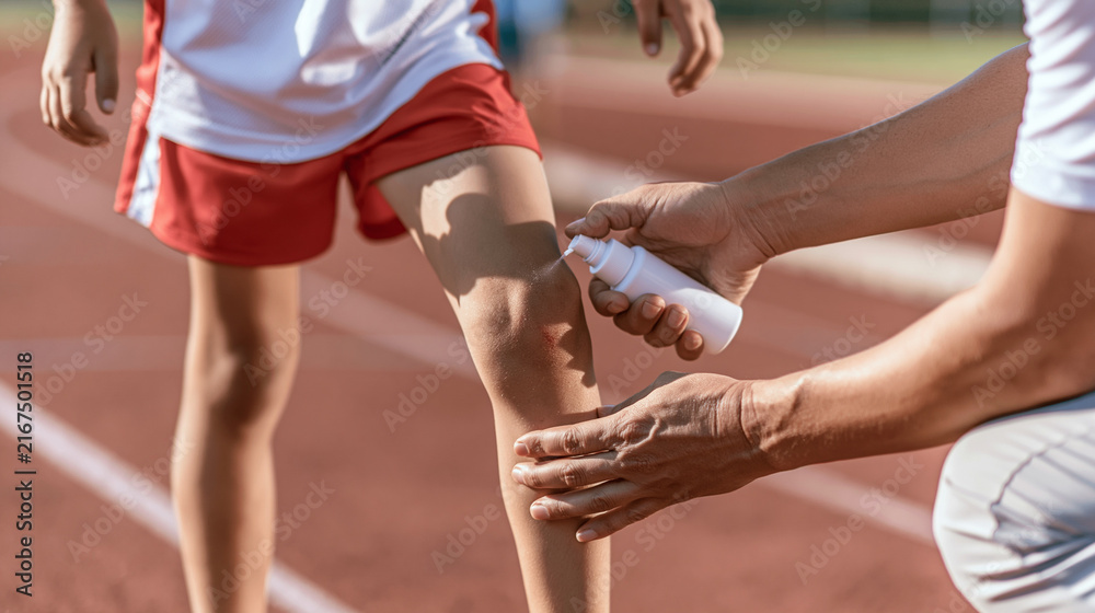Fototapeta premium First aid applying disinfectant spray on child athlete knee injury after falling during running practice on track
