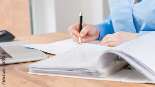 Person writes with a pen in a blue shirt. Laptop rests on a wooden desk. Open notebook lies beside their work. They concentrate fully on the task. This scene suits office or academic settings