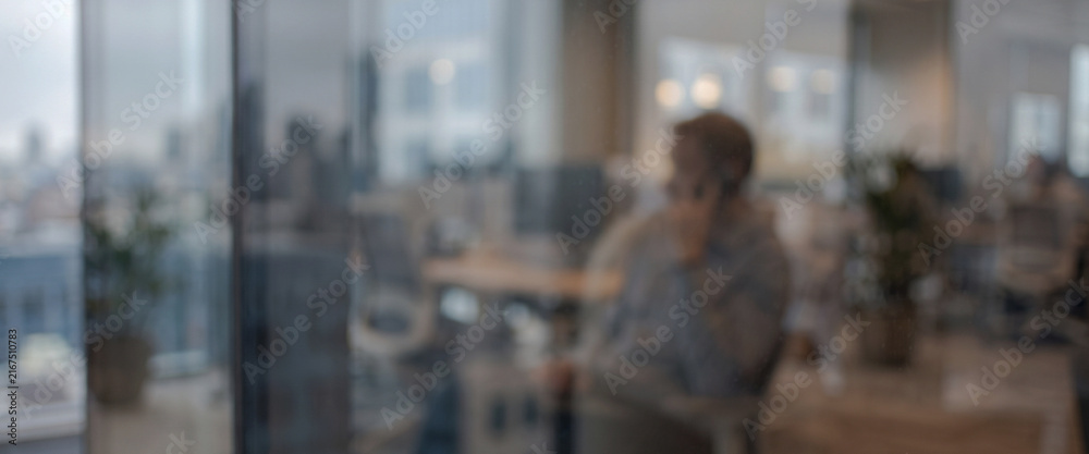 Fototapeta premium A blurred image of a person seated in an office, with modern workspace elements in the background and city views visible through the windows.