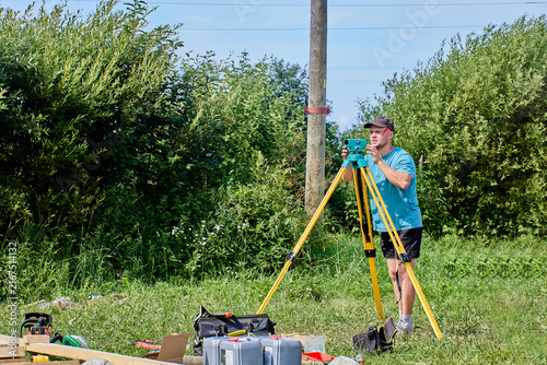 Surveyor operates precision instrument mounted on yellow tripod outdoors, focusing on distant target point.