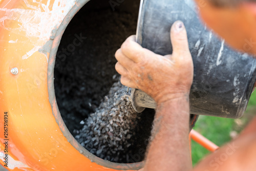 Worker pouring gravel into a concrete mixer. Close-up of hands pouring crushed stone into an orange concrete mixer, symbolizing the construction process and mixture preparation