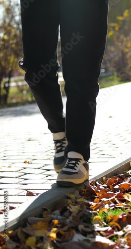 Young woman walks along curb in autumn park, balancing carefully in warm sunlight. Close view of casual clothing, sneakers and fallen leaves, suitable for lifestyle, movement and outdoor content.