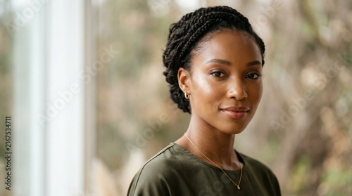 Elegant Black woman with angular face and glowing warm-toned skin in close-up beauty portrait, soft directional window light, olive top, braided updo hairstyle, gold necklace, bokeh greenery