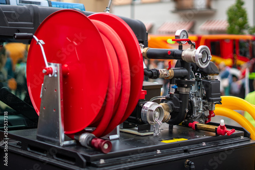 Cuadro en lienzo Fire truck featuring a red fire hose reel in the foreground, water pump unit, pressure gauge, and firefighting equipment, captured in a close-up shot in bright daylight