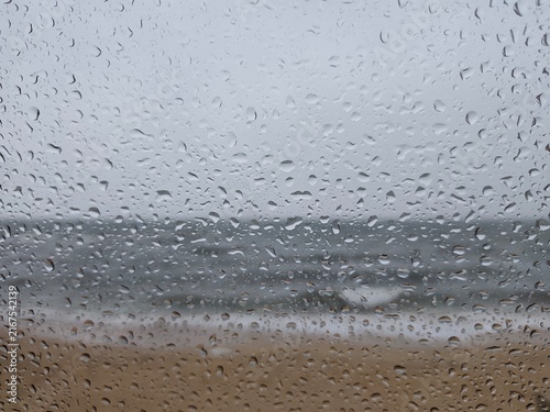 Photography Full frame view of water droplets clinging to a glass window with a blurred view of a stormy sea and beach in the background under an overcast sky